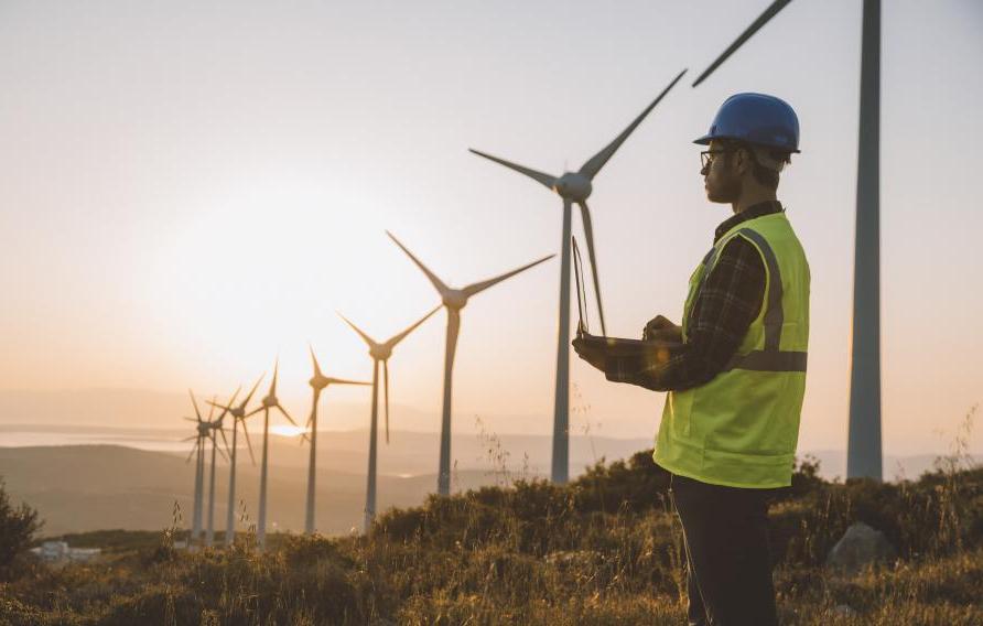 Silhouette of young male engineer holding laptop computer planning and working for the energy industry and standing beside a wind turbines farm power station at sunset time. XXXL size taken with Canon 5D MIV